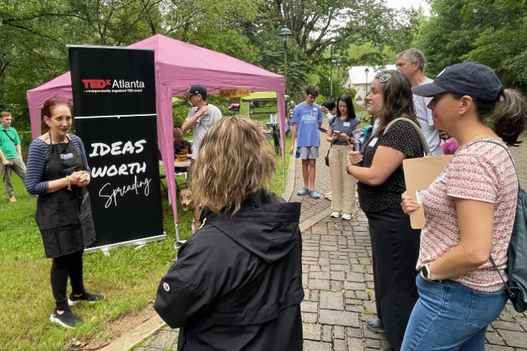 Speaker addresses a small group at an outdoor TEDxAtlanta event with a banner and canopy tent in the background.