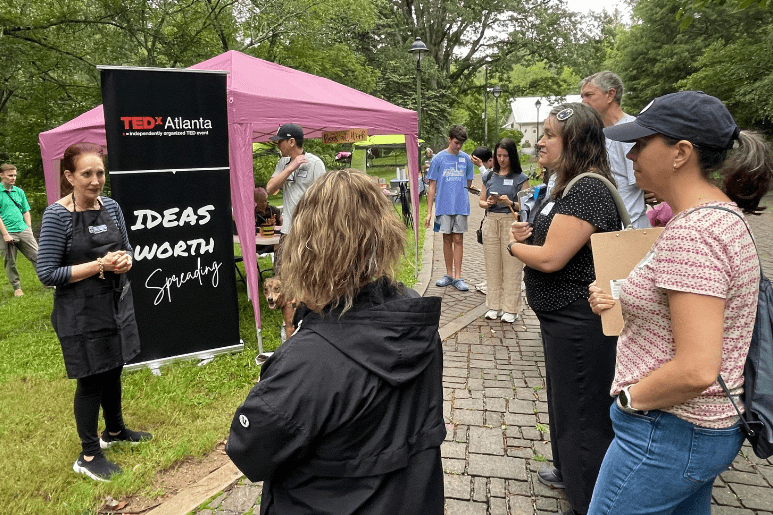 Speaker addresses a small group at an outdoor TEDxAtlanta event with a banner and canopy tent in the background.