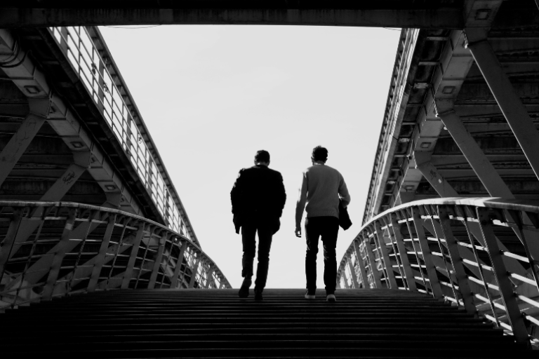 Two people walk side by side up a set of stairs beneath a steel pedestrian bridge, photographed in black and white from behind.